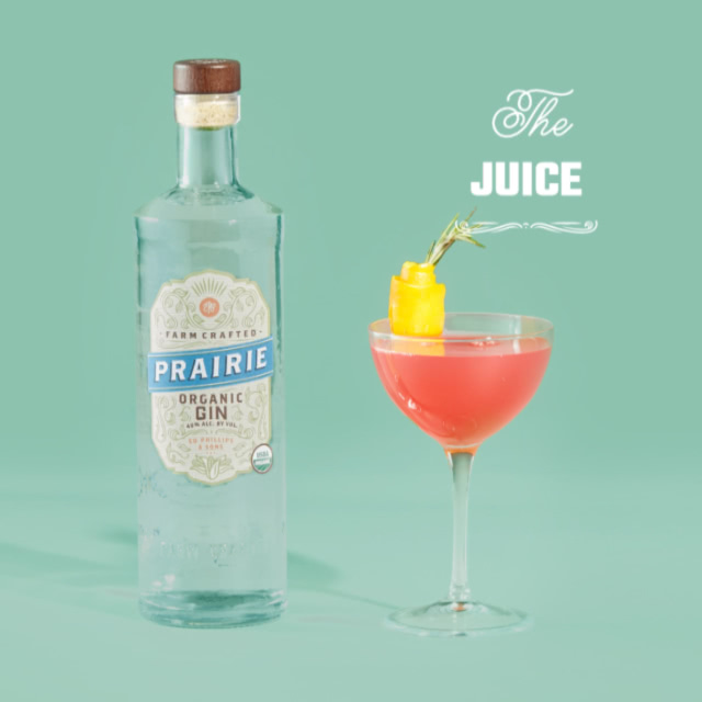 A cocktail glass with a peach colored drink next to a bottle of Prairie Organic Gin